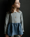 A young girl with long brown hair wearing a striped jacket and plaid pants stands against a dark background.