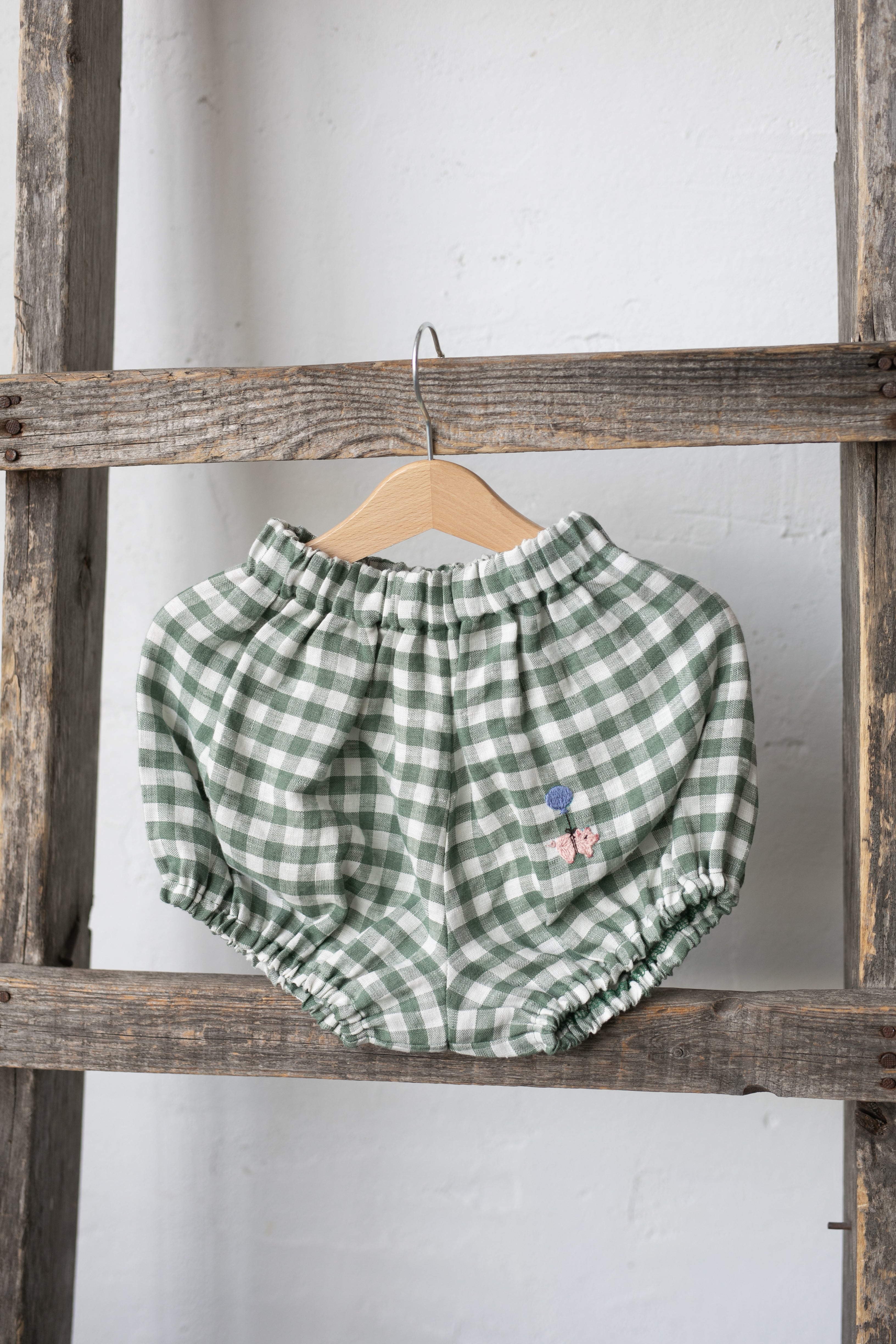 A pair of green and white checkered shorts hanging on a wooden hanger against a white wall.