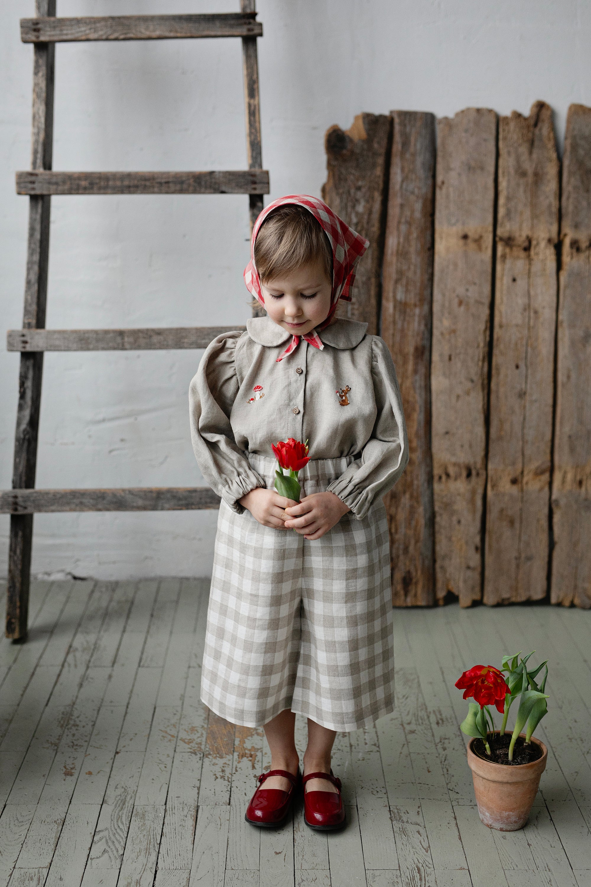 A young girl in a checkered dress and red shoes holds a red flower in front of a wooden ladder and a potted plant.
