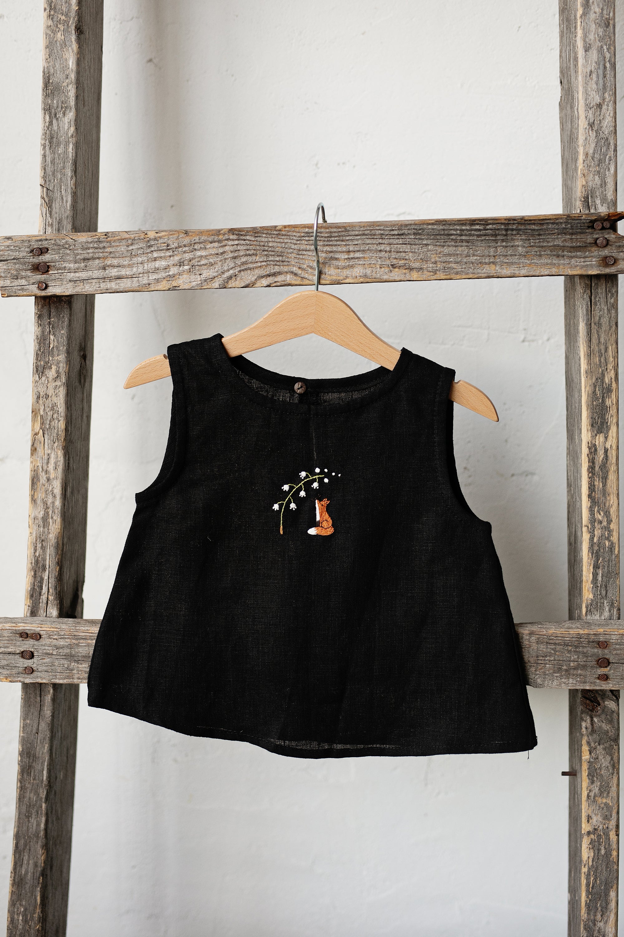 A black sleeveless dress with a fox embroidered on the front is hanging on a wooden hanger.