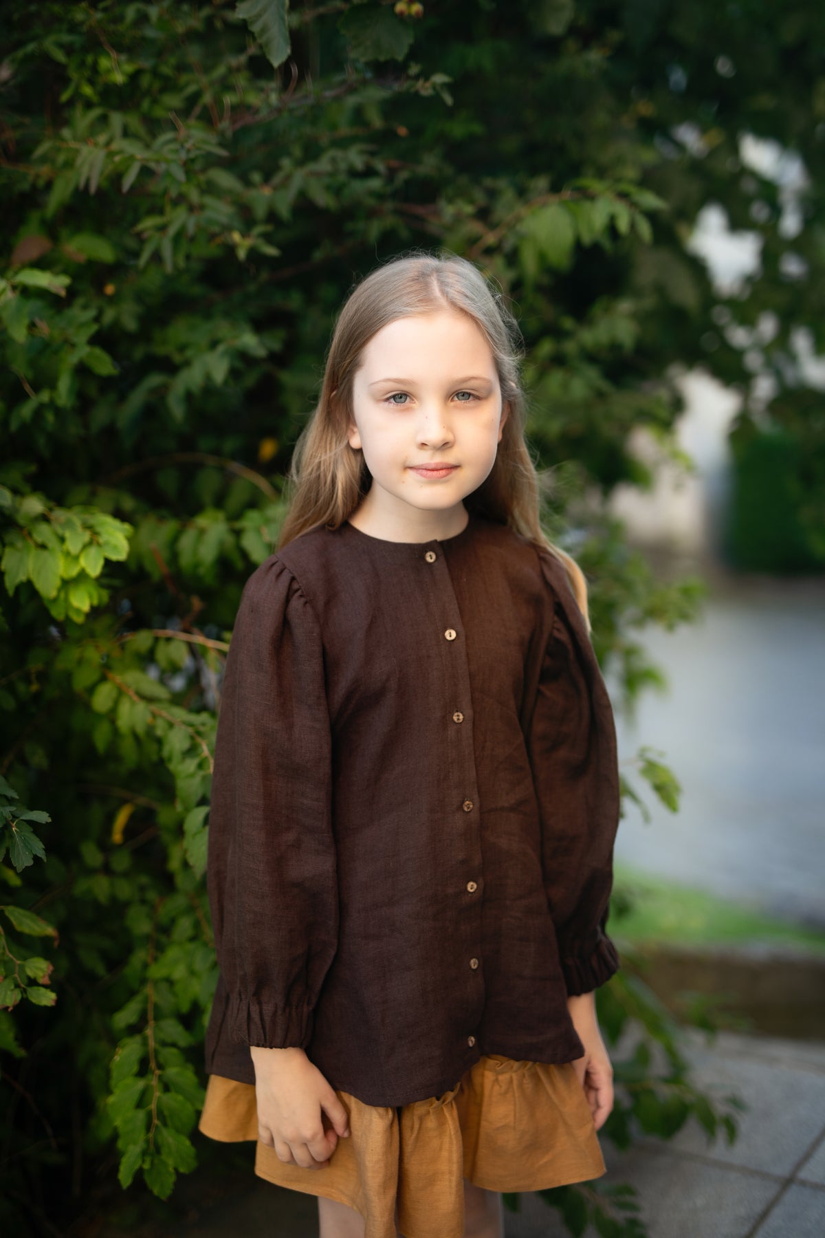 A young girl with long blonde hair stands in front of a lush green bush, wearing a brown blouse with puffed sleeves and a brown skirt.