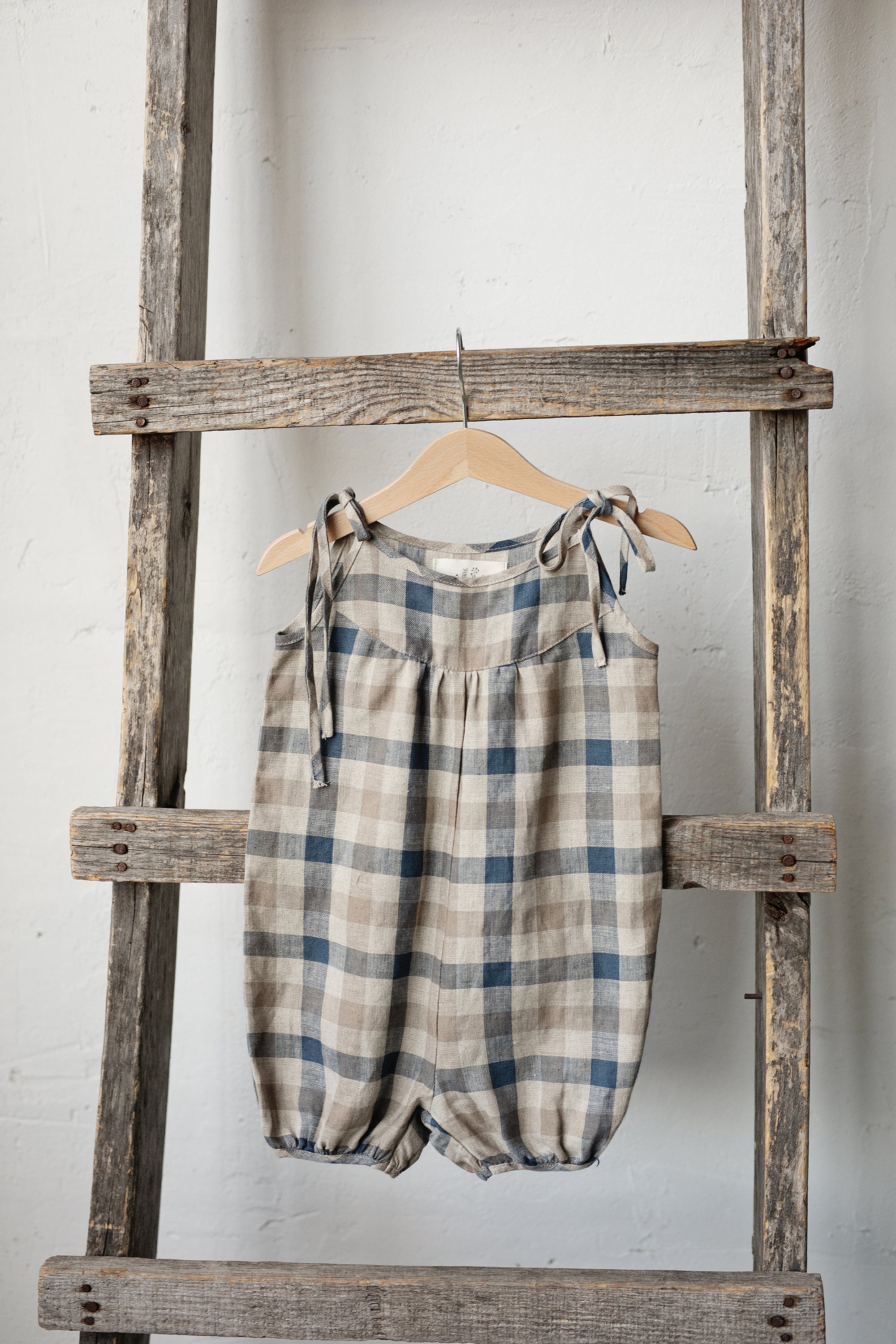 A plaid onesie is hanging on a wooden ladder against a white wall.