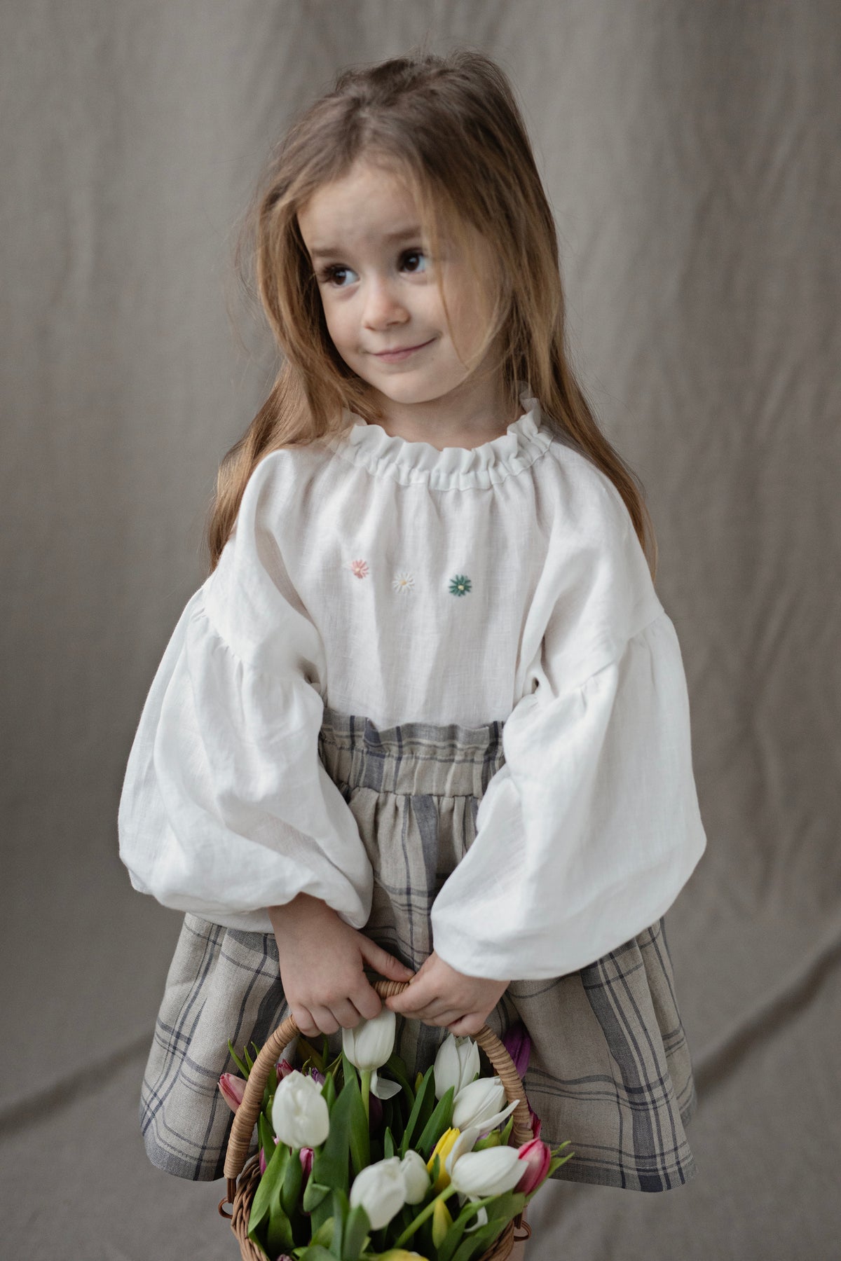 A young girl with long brown hair is holding a basket filled with white tulips. She is wearing a white blouse with green and pink flowers on it.