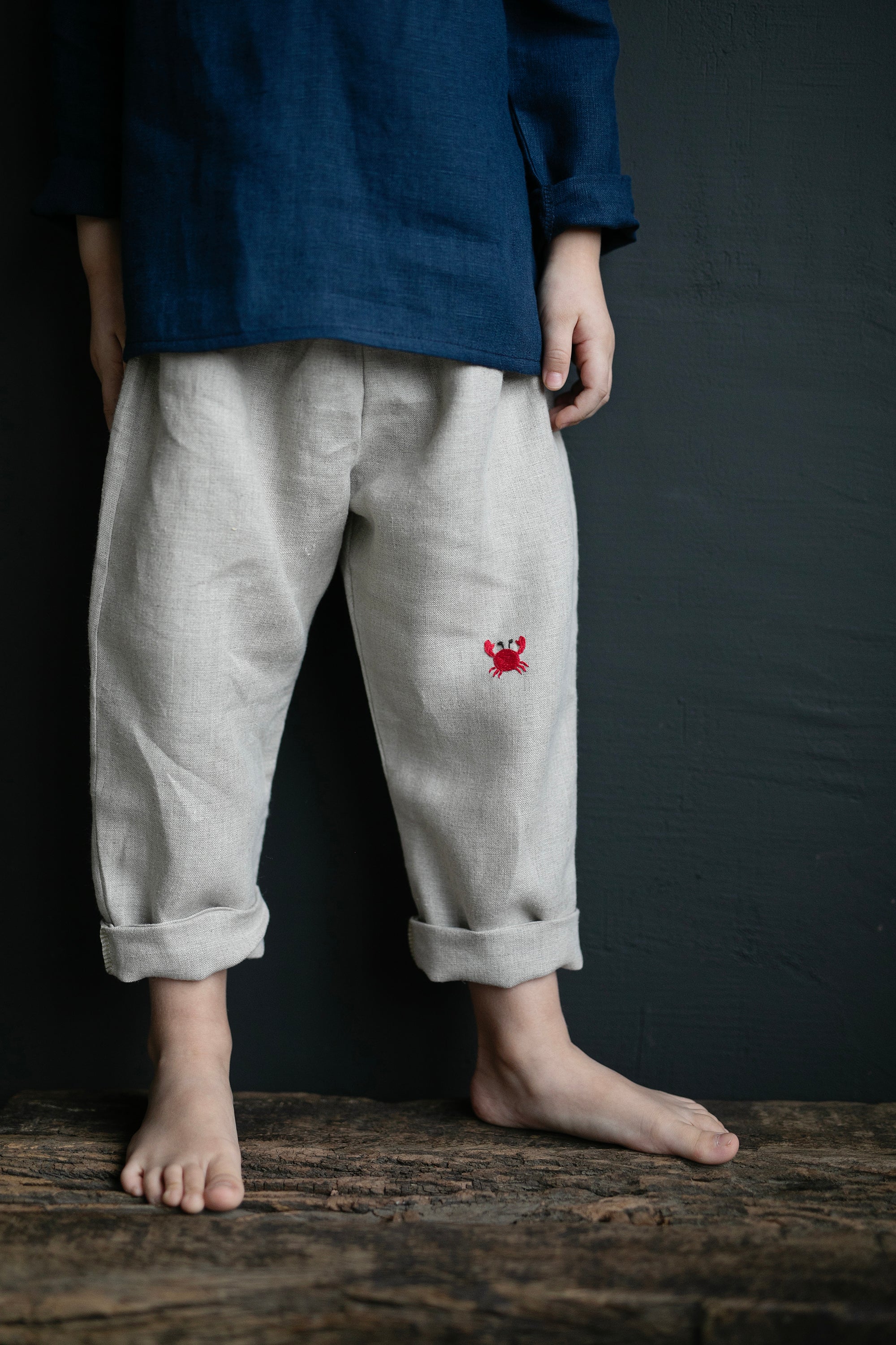 A person wearing a blue shirt and gray pants with a red crab embroidered on the front, standing on a wooden surface against a dark background.
