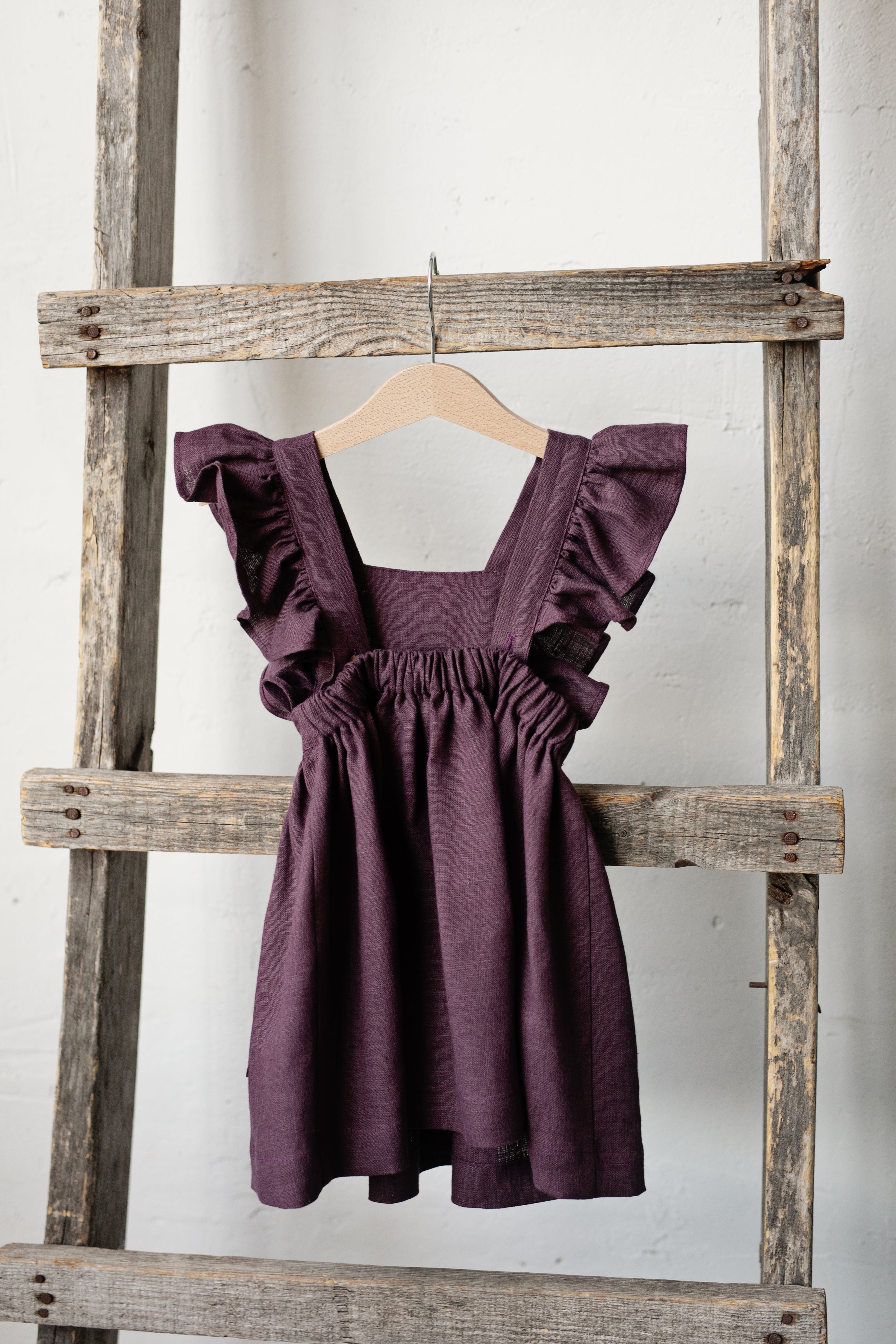 A purple dress with ruffled details is hanging on a wooden ladder against a white wall.