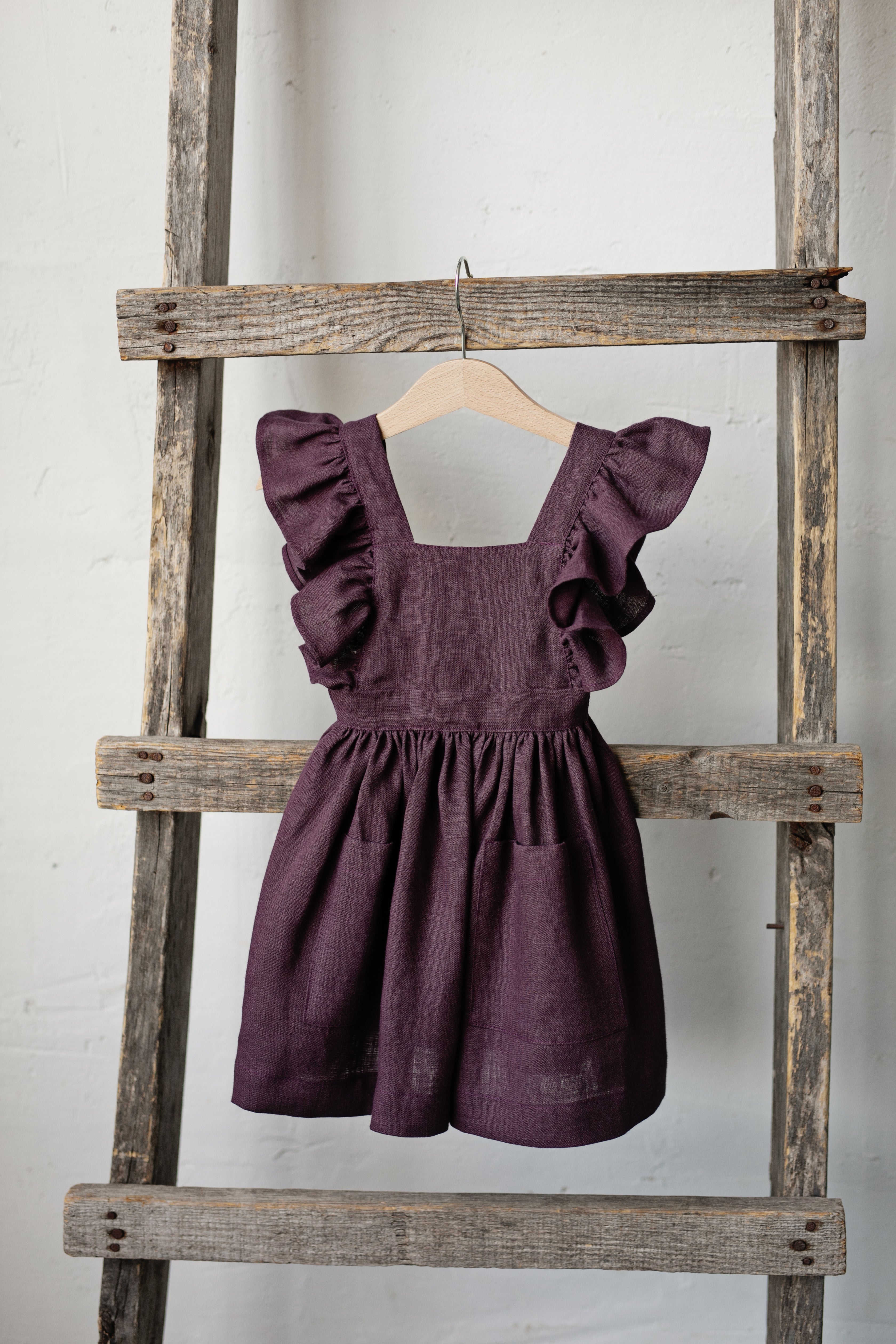 A purple dress with ruffled sleeves and a ruffled hem is hanging on a wooden ladder against a white wall.