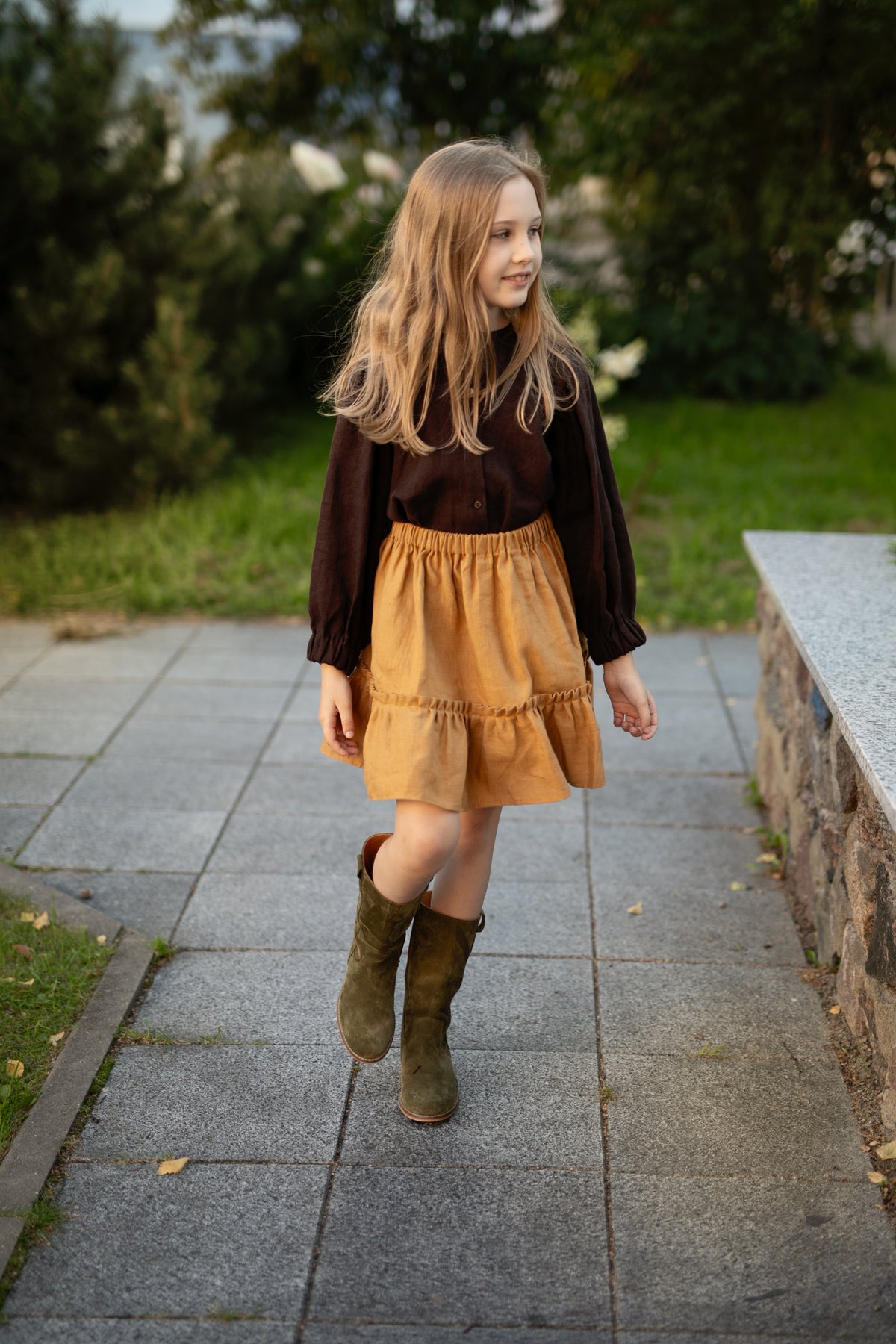 A young girl with long blonde hair is walking on a sidewalk, wearing a brown sweater, a yellow skirt, and green boots.