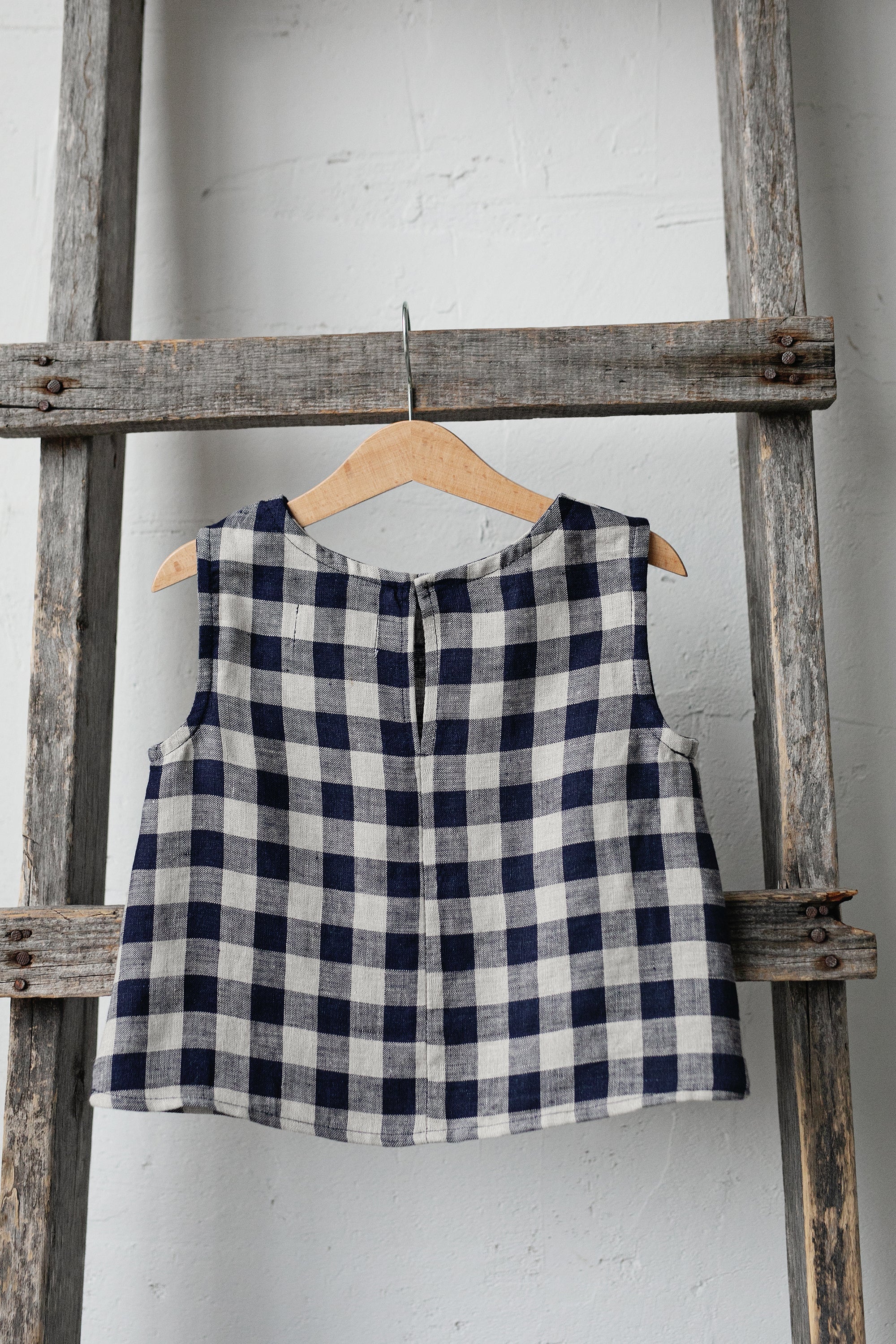 A blue and white checkered dress is hanging on a wooden ladder against a white wall.