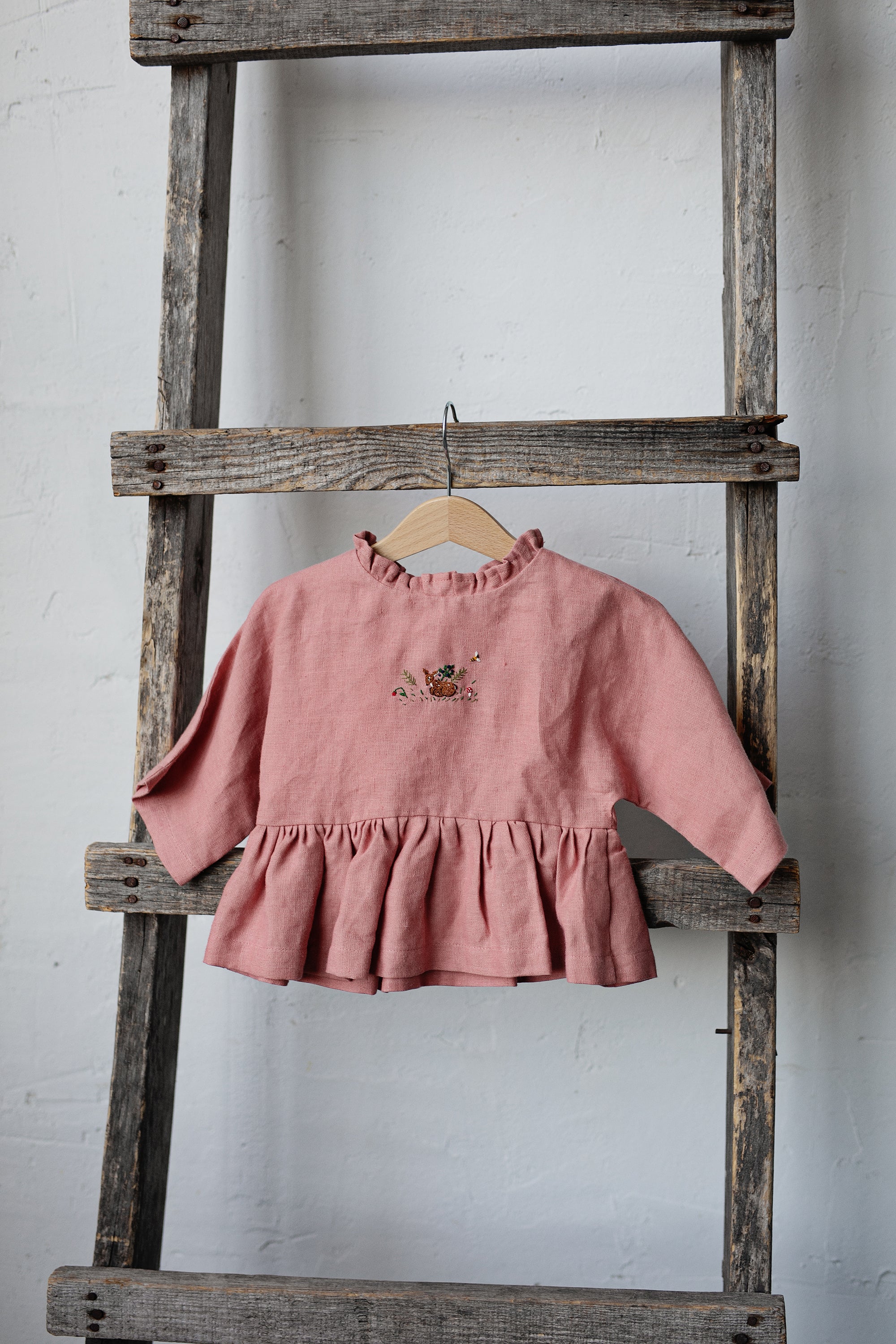 A pink ruffled dress is hanging on a wooden ladder against a white wall.