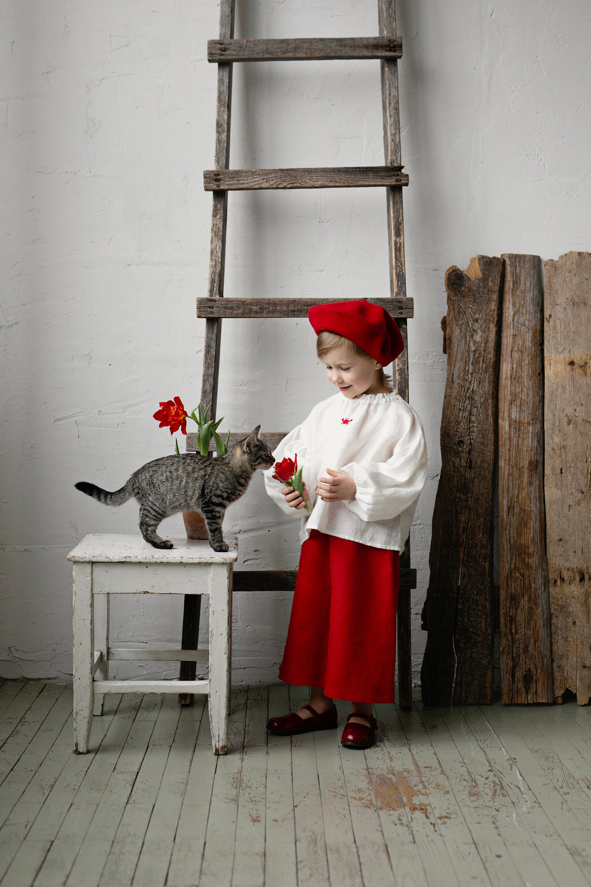 A young girl in a red hat and white blouse stands next to a gray cat, holding a flower, with a wooden ladder and a wooden fence in the background.