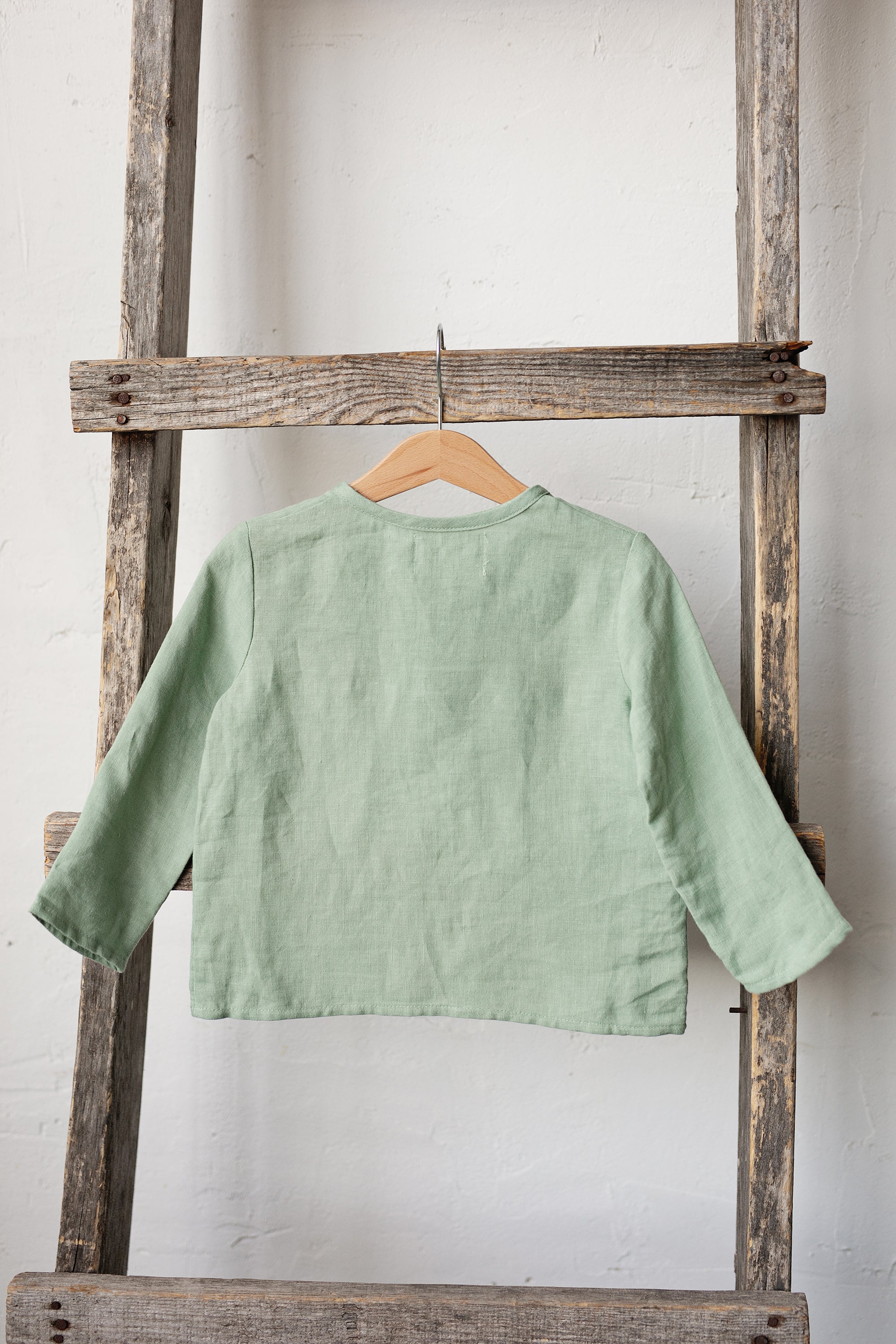 A light green, long-sleeved shirt is hanging on a wooden ladder against a white wall.