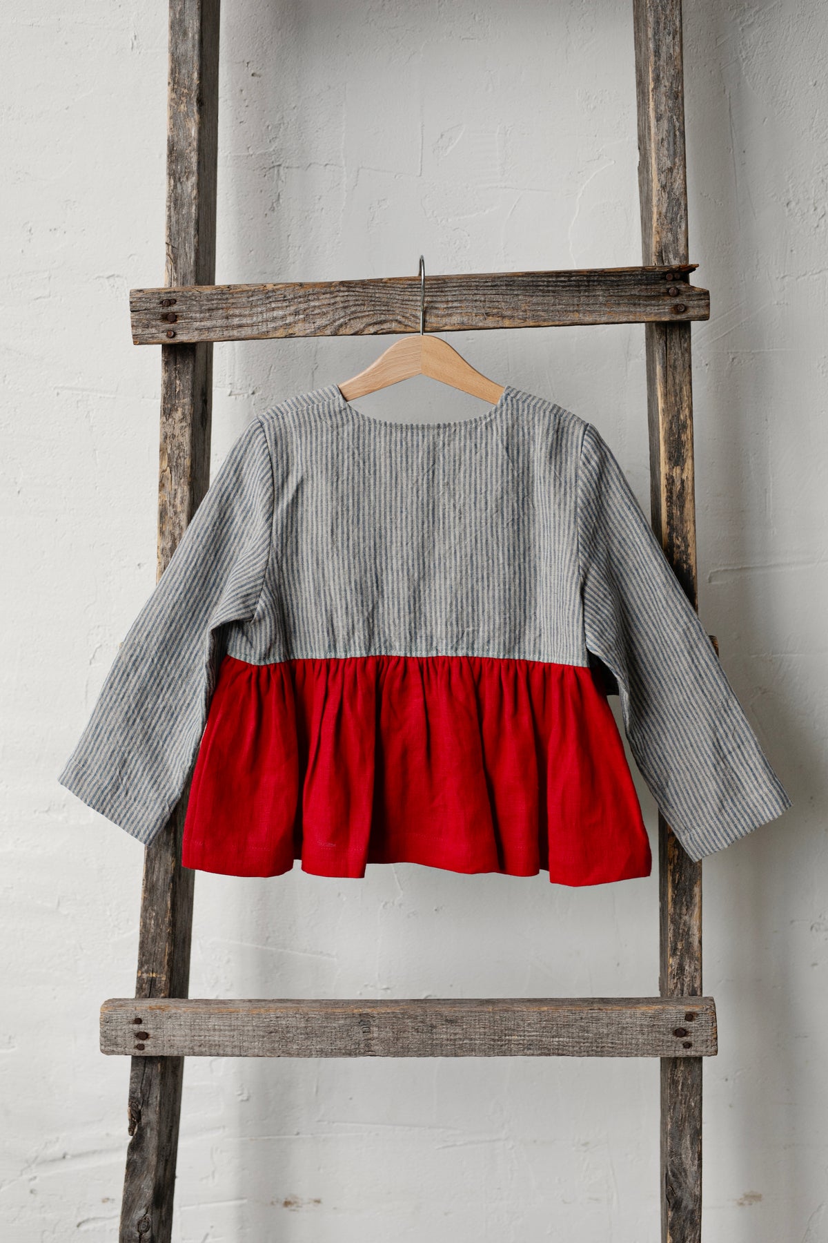 A gray and red striped blouse is hanging on a wooden ladder against a white wall.