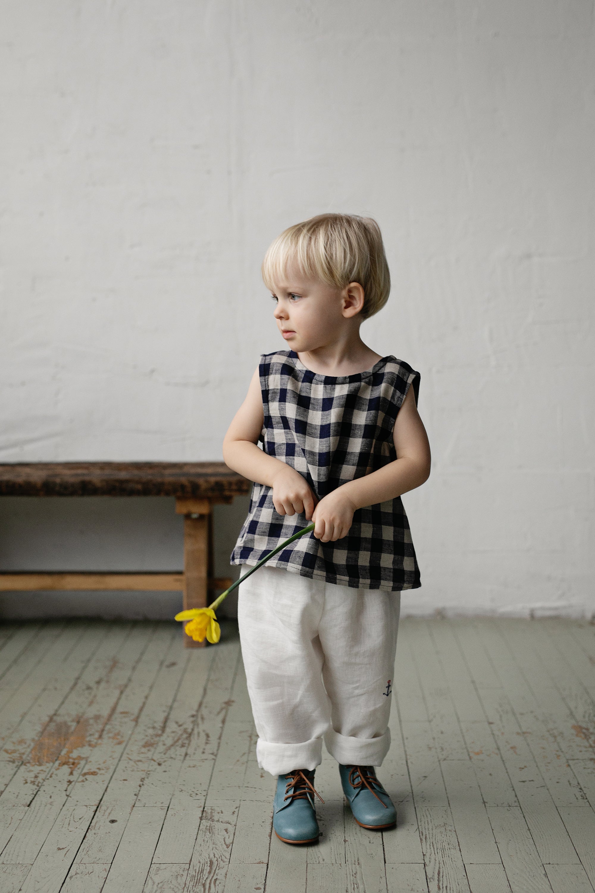 A young child in a black and white checkered shirt and white pants stands in a room, holding a yellow flower in their hand.