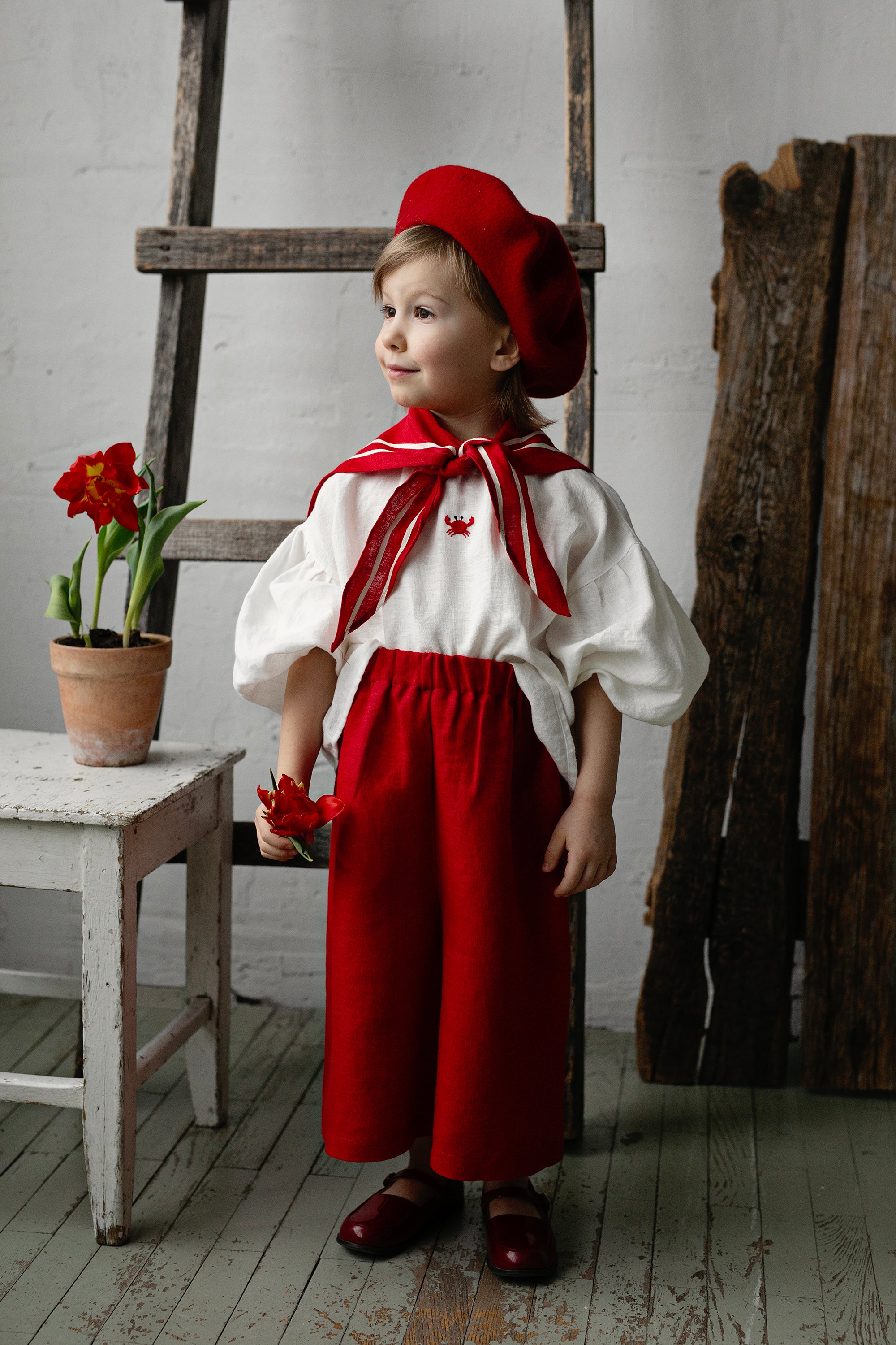 A young child dressed in a red outfit and hat stands in a room with a wooden ladder and a potted plant.