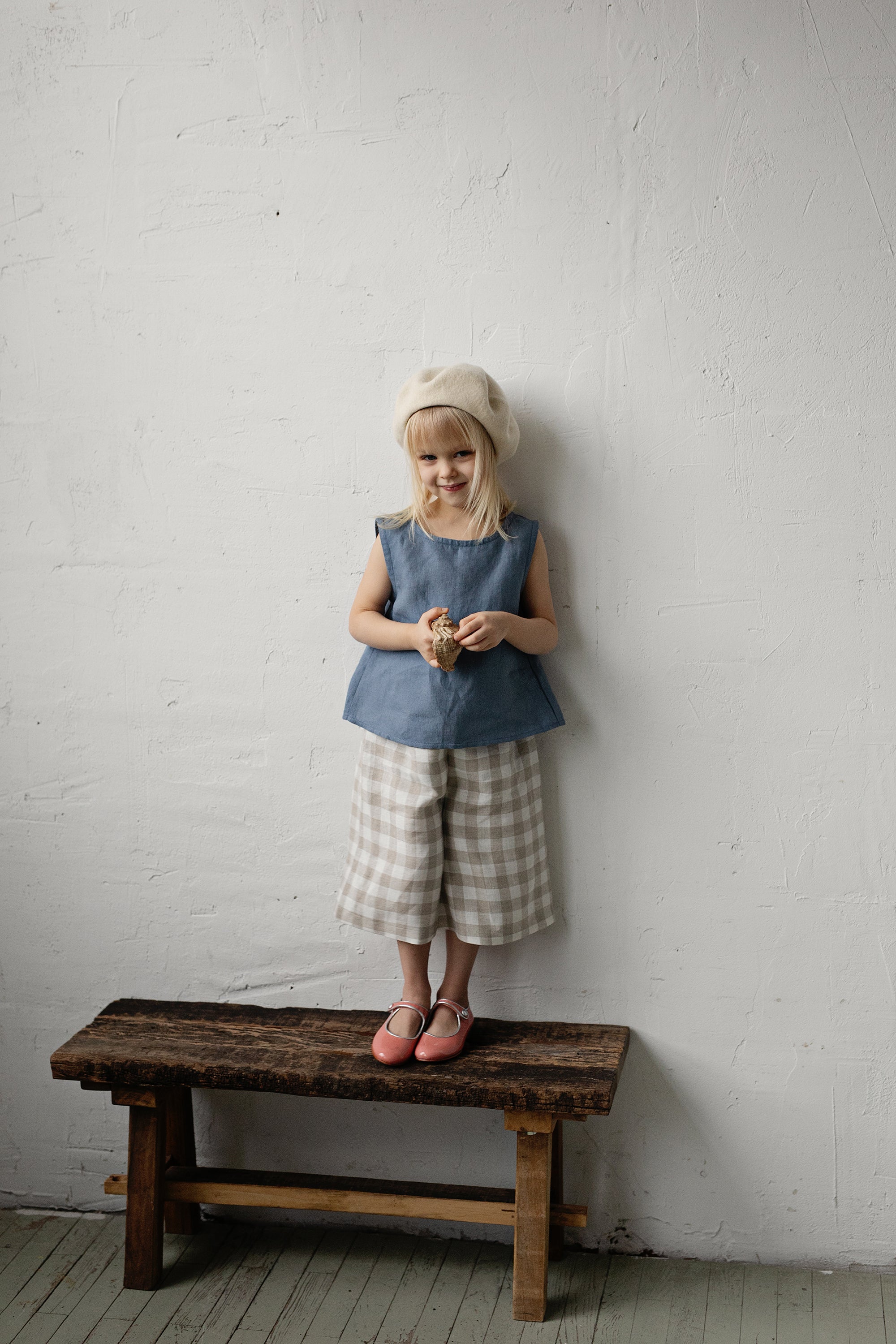 A young girl with blonde hair is standing on a wooden bench, wearing a blue top and plaid shorts, and holding a small object in her hands.
