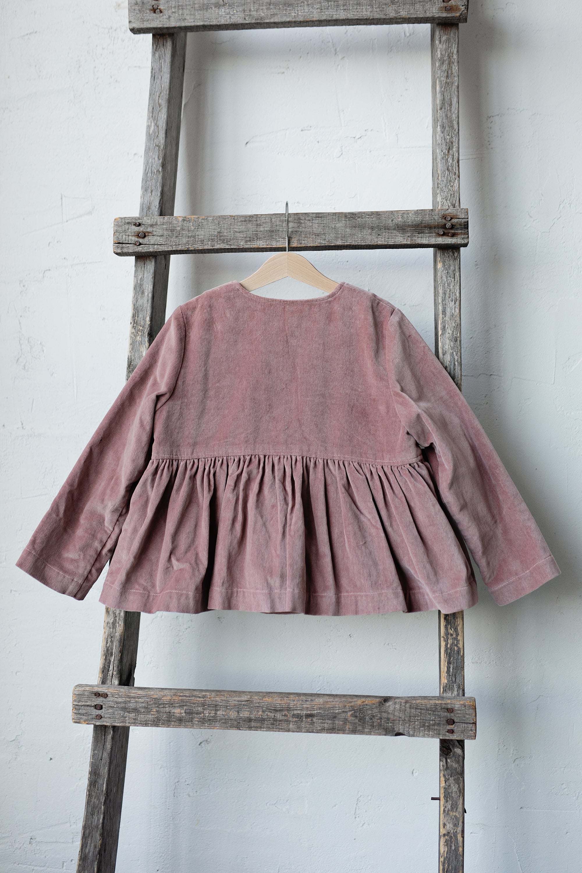 A pink ruffled jacket is hanging on a wooden ladder against a white wall.