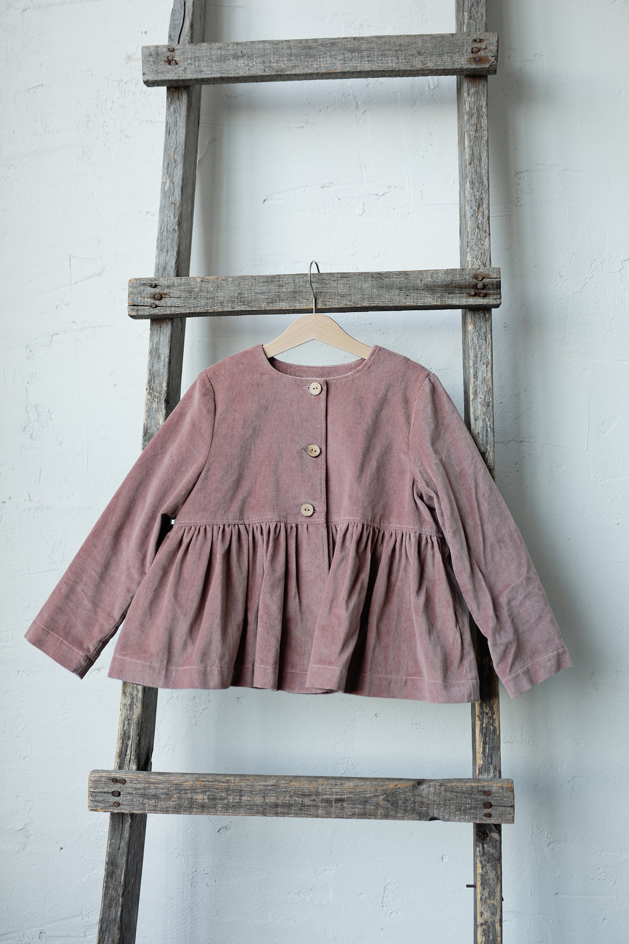 A pink ruffled jacket is hanging on a wooden ladder against a white wall.