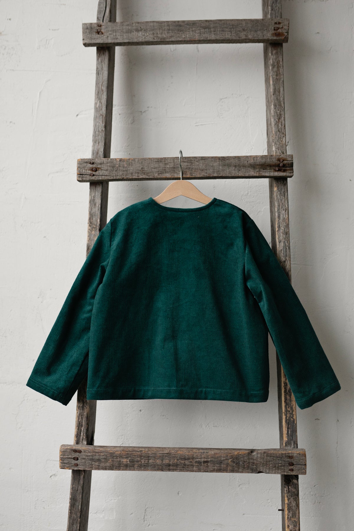 A dark green long-sleeved shirt is hanging on a wooden ladder against a white wall.
