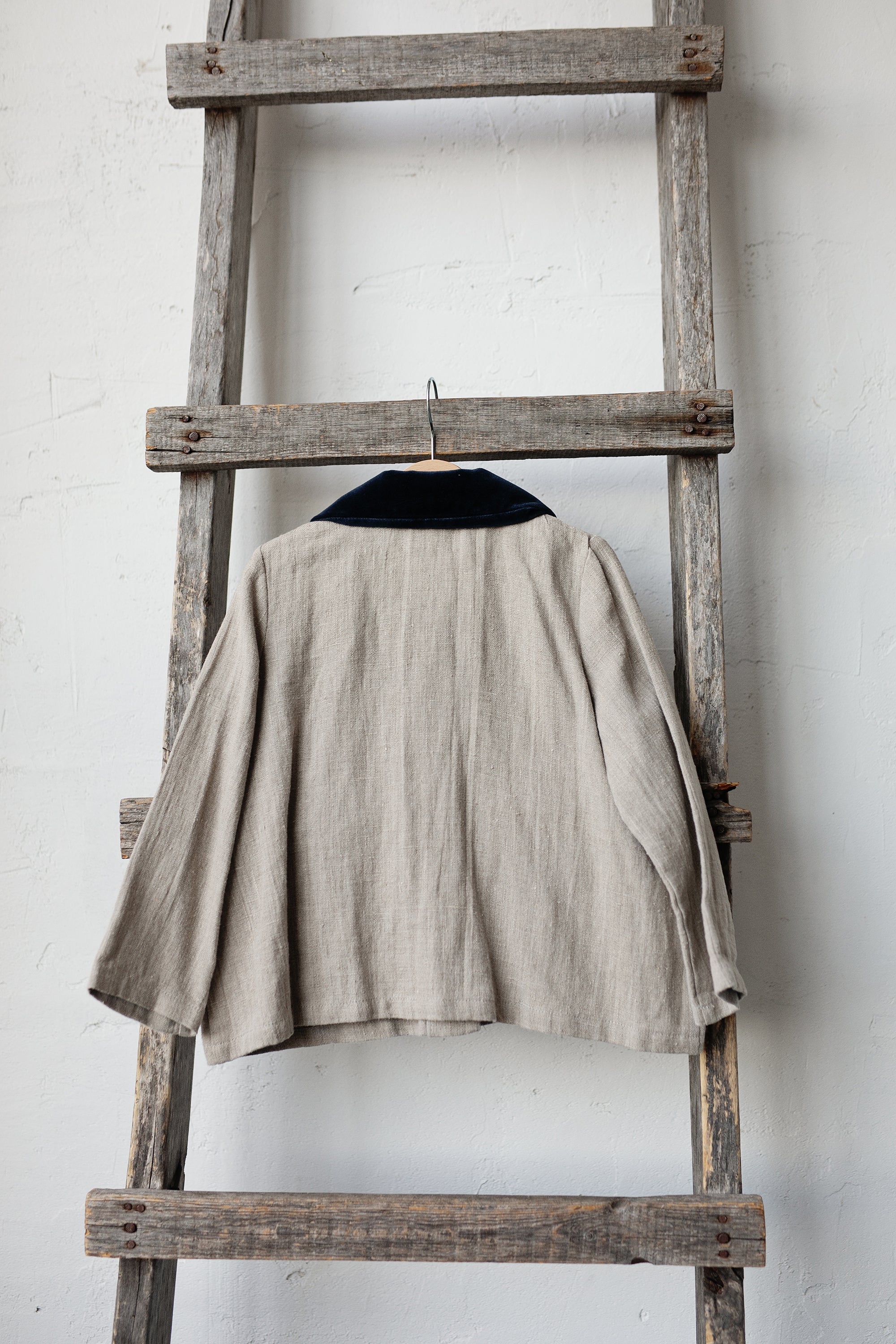 A beige jacket with a black collar is hanging on a wooden ladder against a white wall.