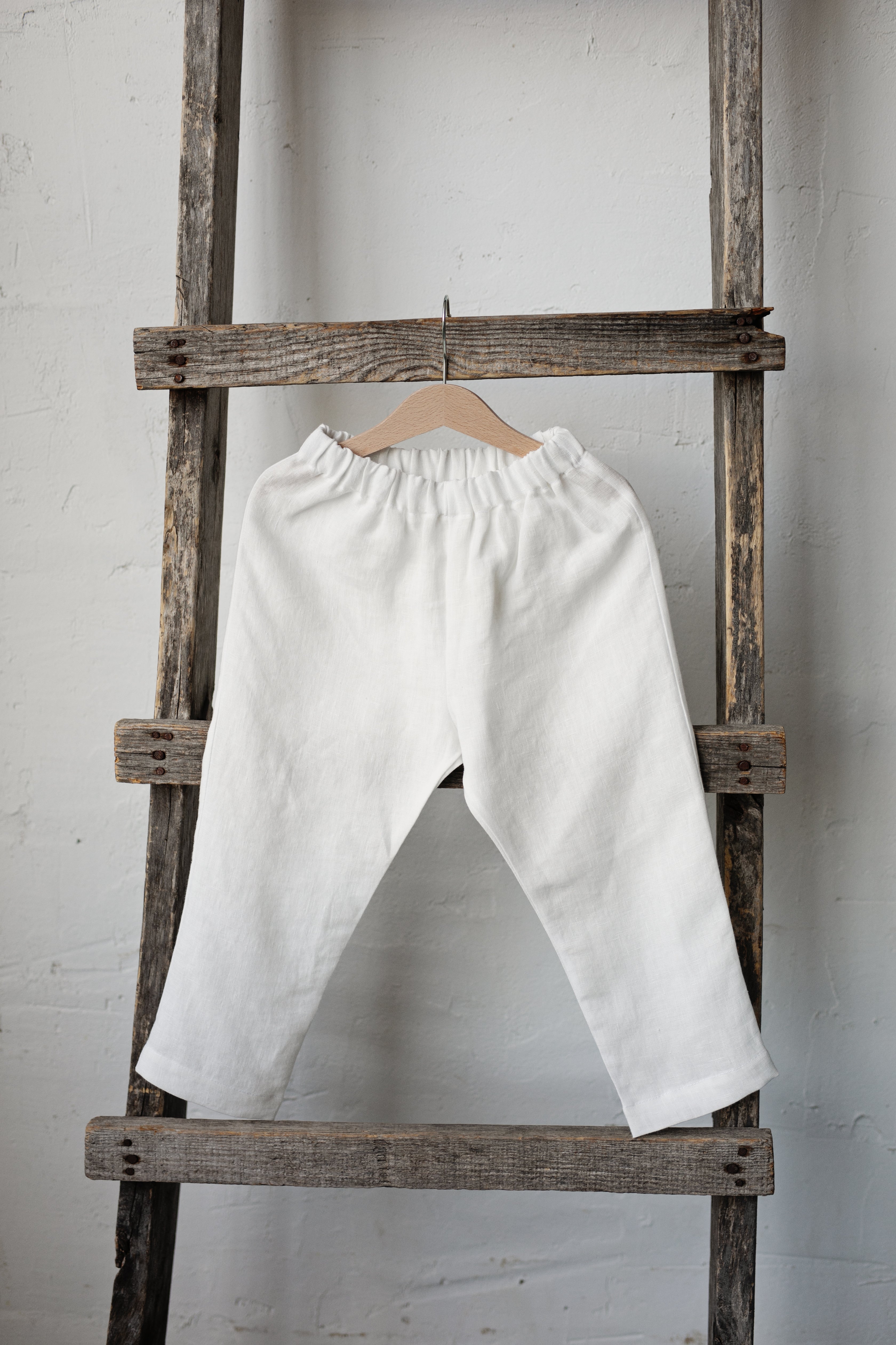 A pair of white pants hanging on a wooden ladder against a white wall.