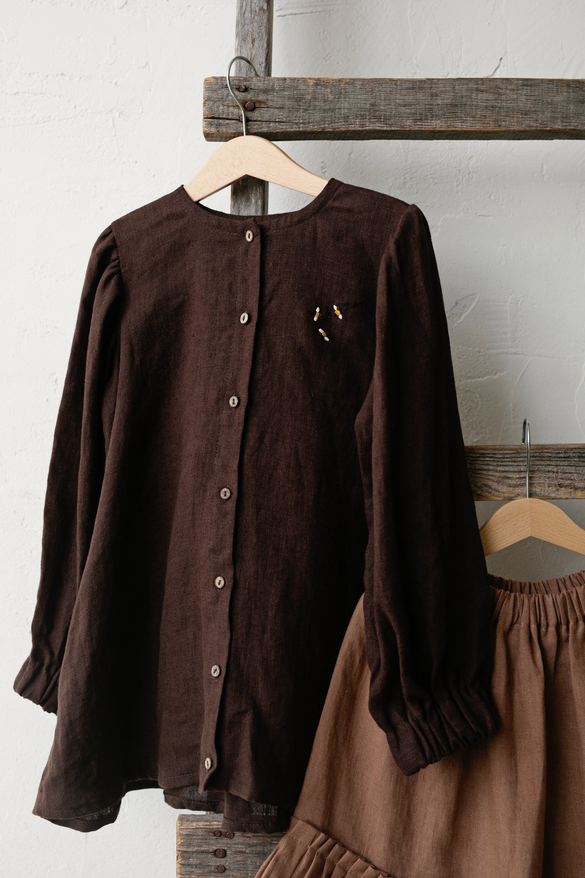 A brown button-up blouse and a brown skirt are hanging on a wooden hanger against a white wall.
