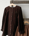 A brown button-up blouse and a brown skirt are hanging on a wooden hanger against a white wall.