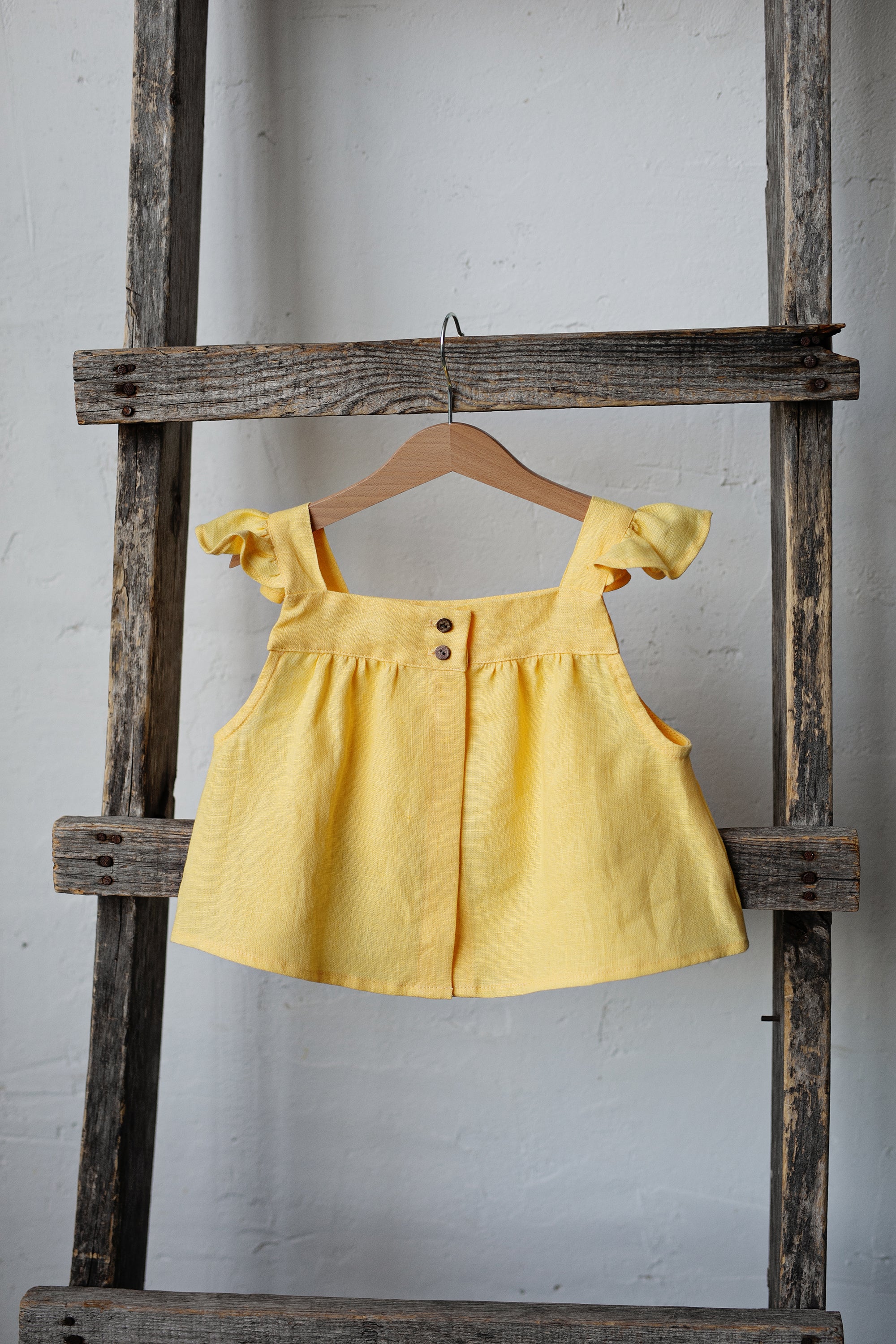 A yellow dress with ruffled sleeves is hanging on a wooden ladder against a white wall.