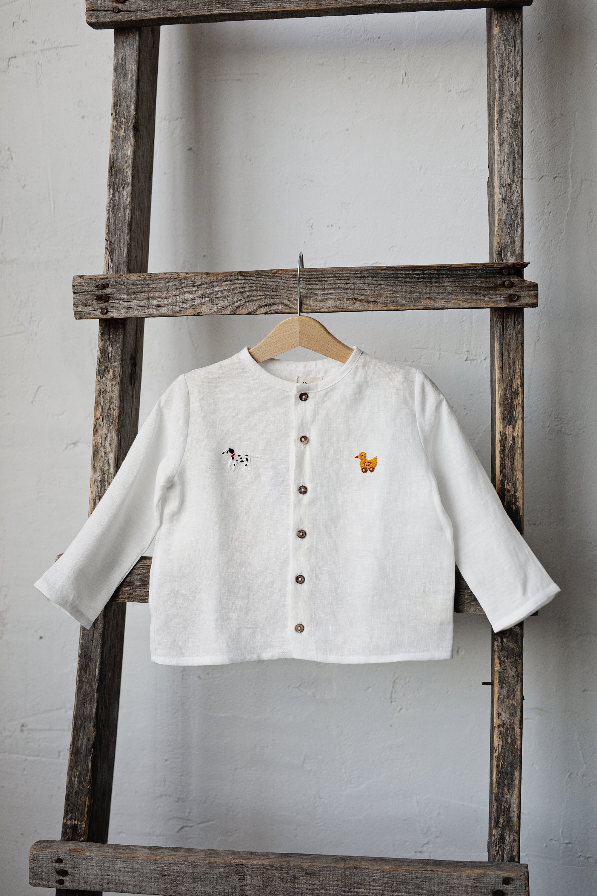 A white button-down shirt with a small embroidered animal design on the left side is hanging on a wooden ladder against a white wall.