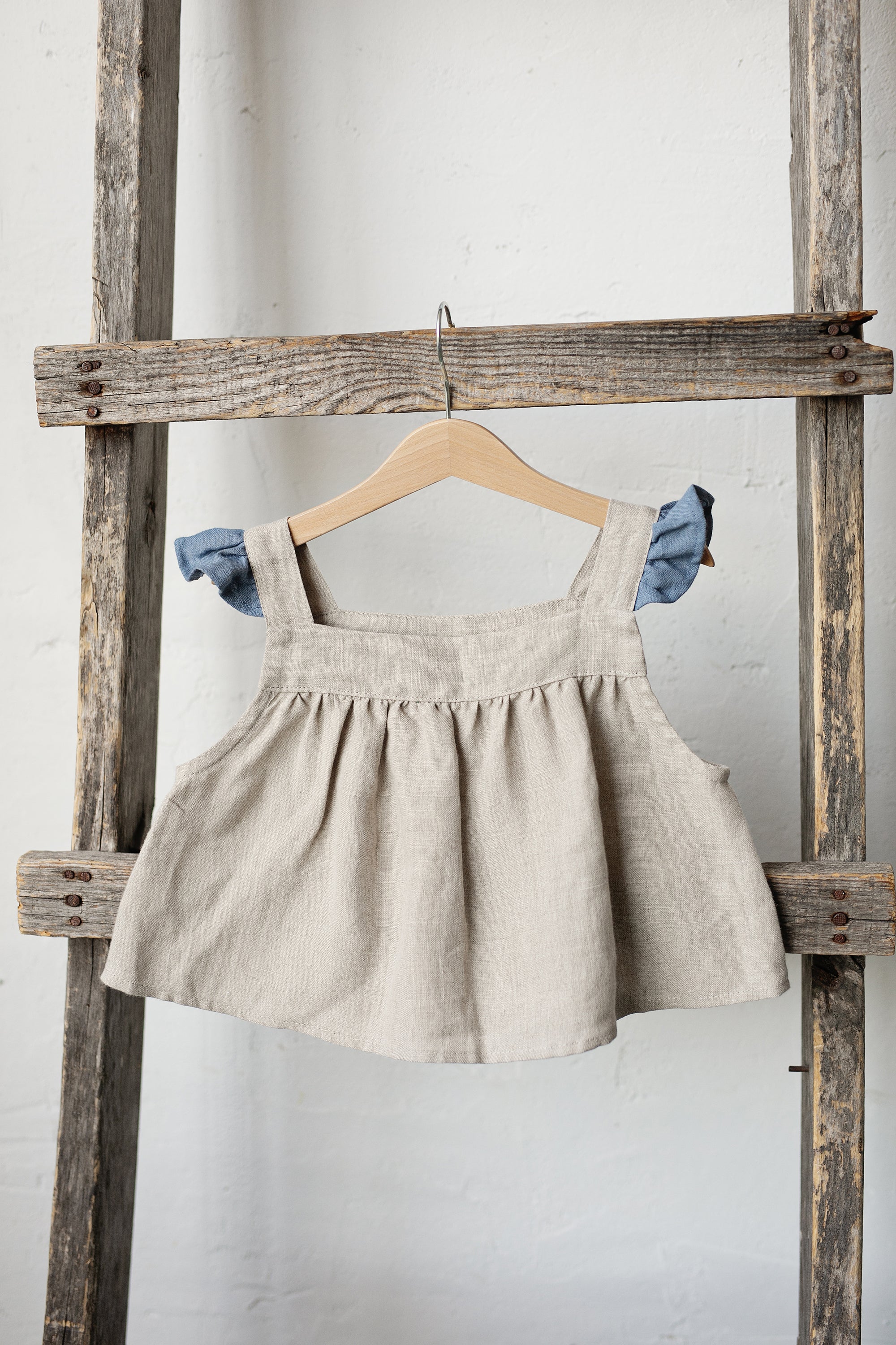 A beige dress with blue accents hangs on a wooden ladder against a white wall.