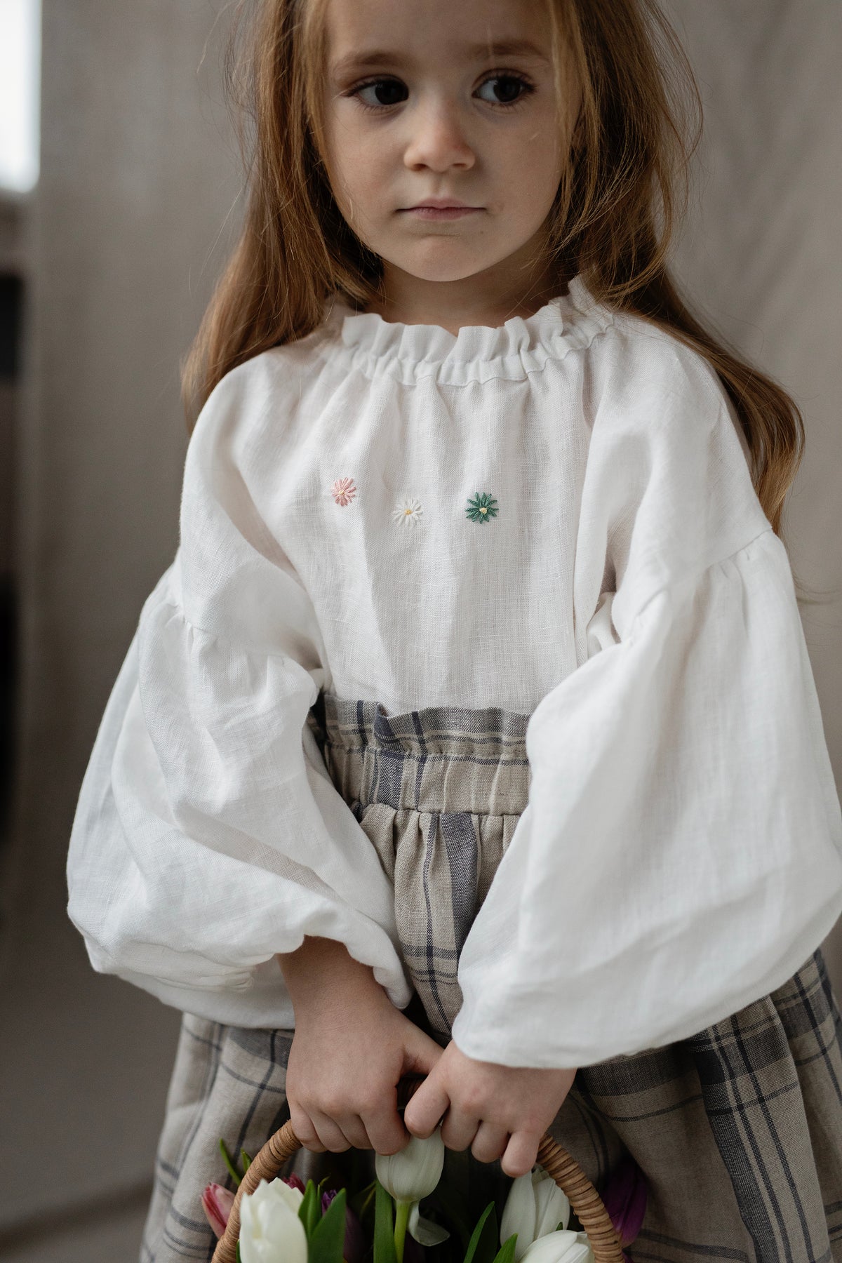 A young girl with long brown hair wearing a white blouse with embroidered flowers and a gray skirt, holding a basket of white flowers.