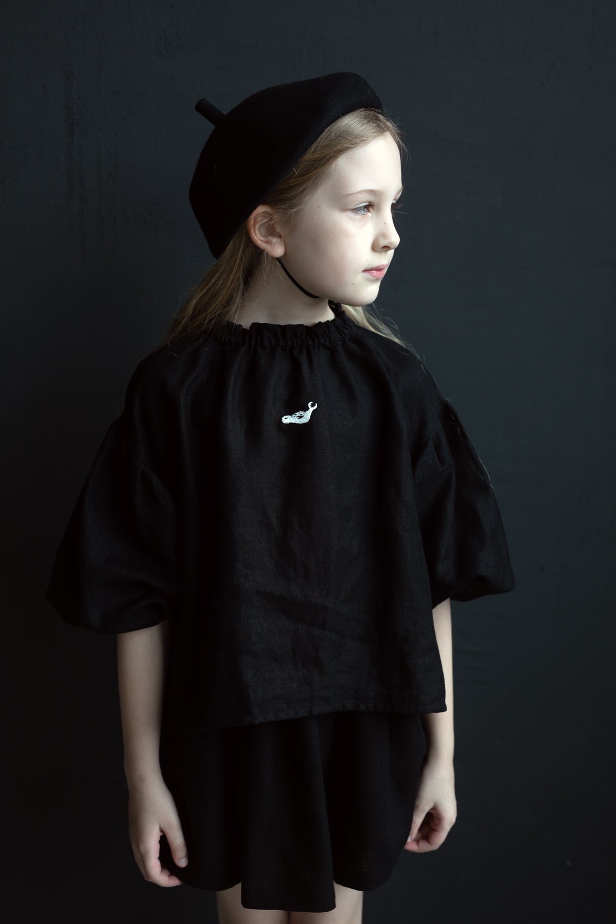 A young girl wearing a black outfit and hat stands against a dark background.