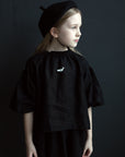 A young girl wearing a black outfit and hat stands against a dark background.