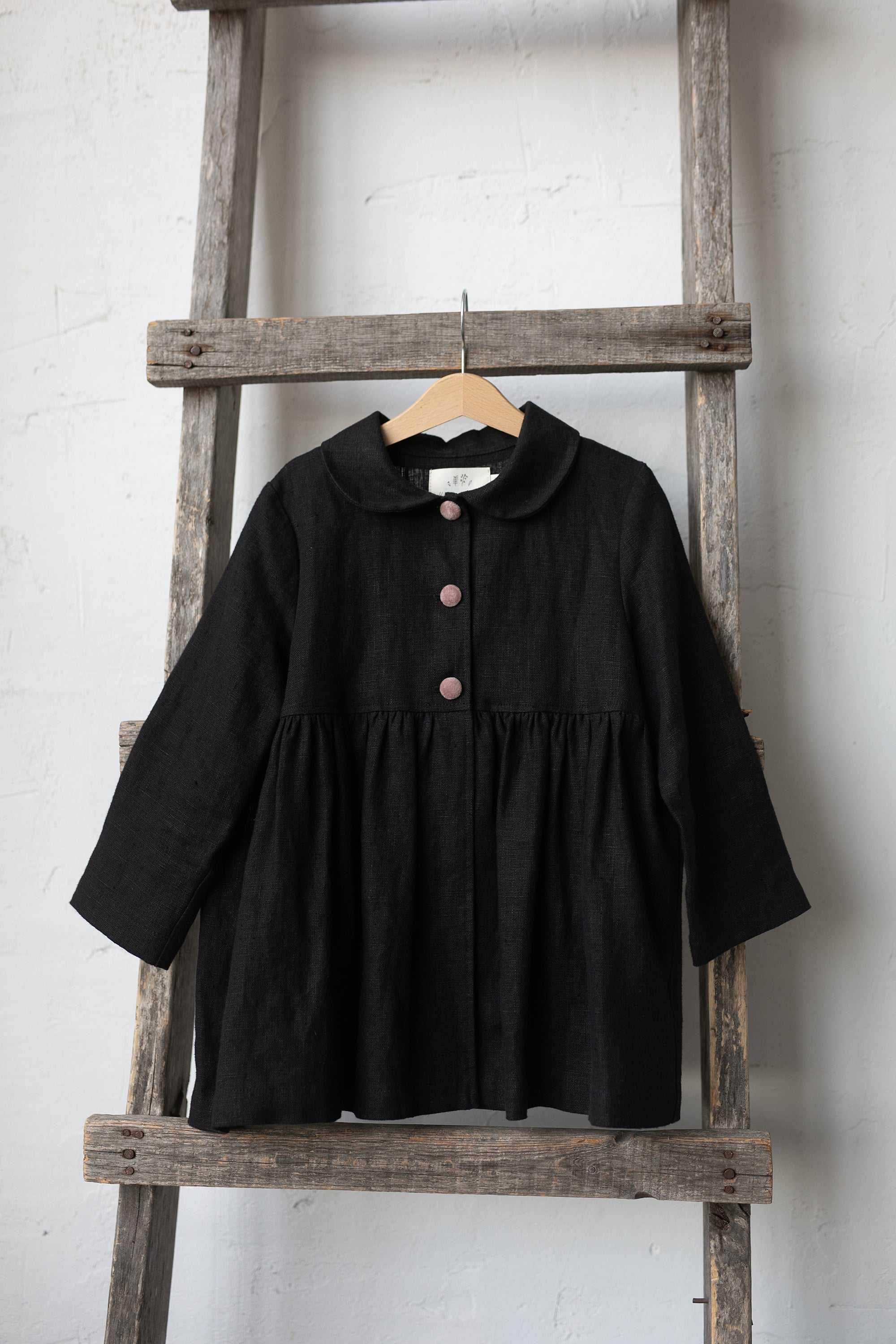 A black, button-up jacket is hanging on a wooden ladder against a white wall.