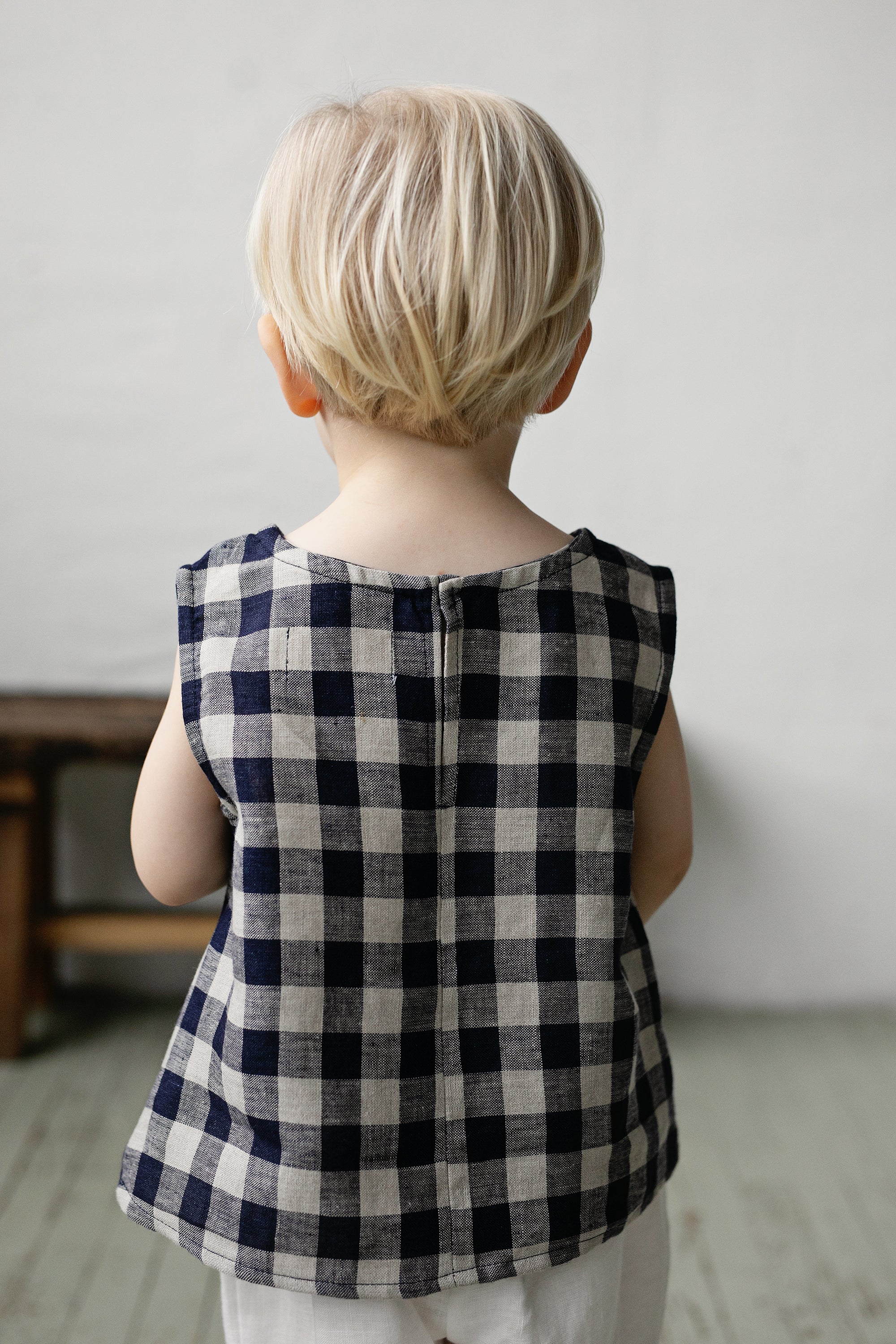 A young child with blonde hair is standing in front of a plain white wall, wearing a blue and white checkered shirt.