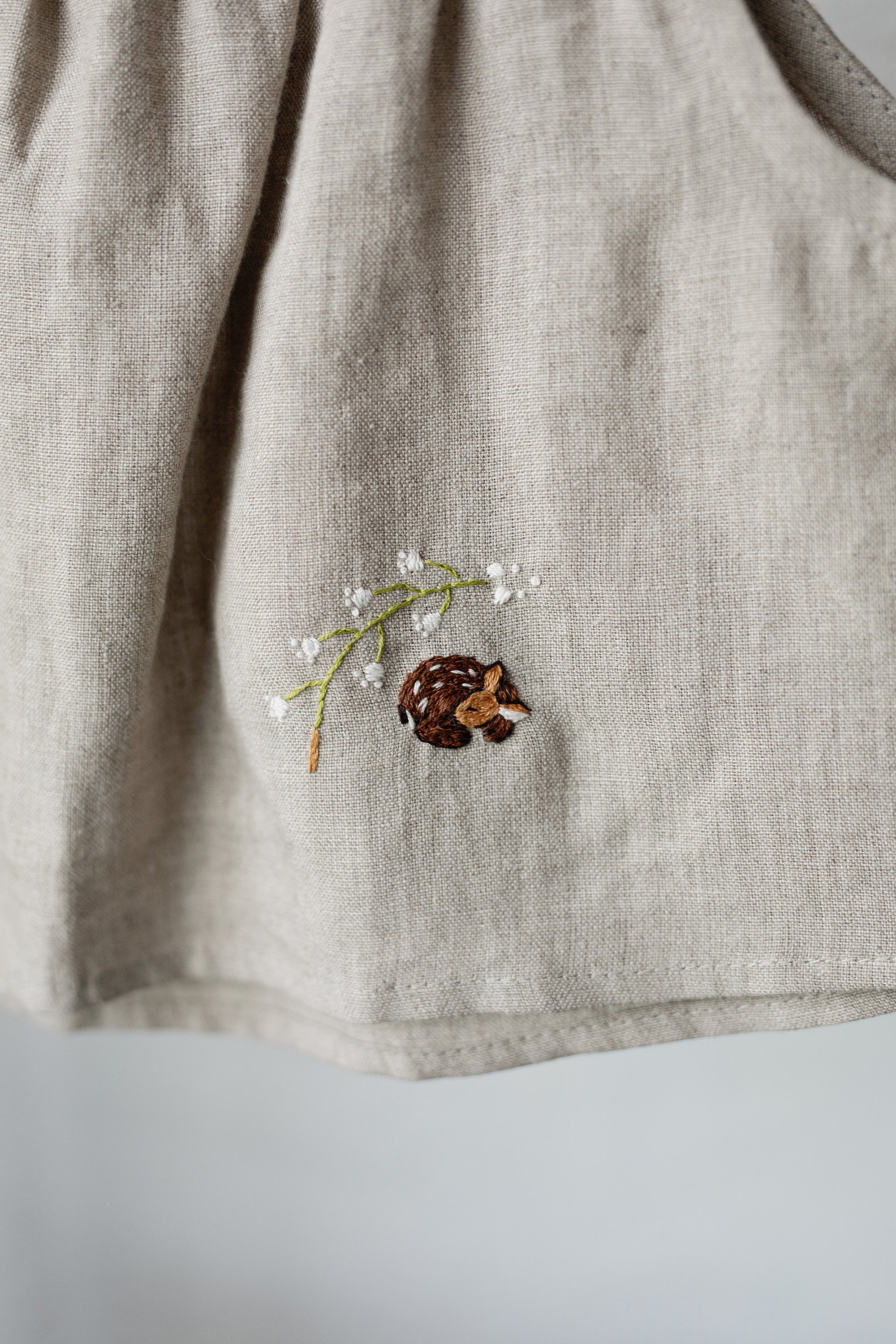 a close-up view of a beige fabric with a small embroidered design featuring a pine cone and a small animal, likely a hedgehog, on it.